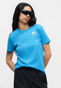 Γυναίκα με μακριά σκούρα μαλλιά που φοράει μαύρα αθλητικά γυαλιά ηλίου, μπλε μπλουζάκι Nike και μαύρο παντελόνι, στέκεται μπροστά σε απλό φωτεινό φόντο.