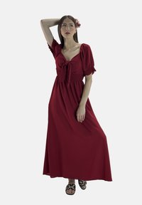 Rotes, knöchellanges Kleid mit kurzen Puffärmelchen und einem Bindeband-Detail; mit gerüschtem Taillenbund. Kombiniert mit schwarzen Sandalen mit geometrischem Muster.