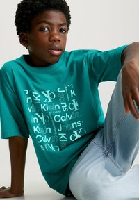 Camiseta de algodón en color azul verde con un diseño de texto de logo repetido en blanco y azul claro. Modelo combinado con jeans de mezclilla azul claro, sentado de manera casual.