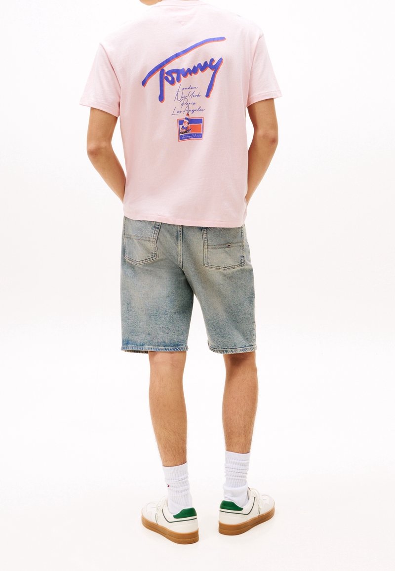 Persona vestida con una camiseta gráfica de color rosa claro y pantalones cortos de mezclilla azul claro, con zapatillas blancas y calcetines blancos, frente a un fondo blanco.