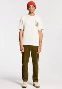 Fehér pamut póló grafikai mintával a bal mellrészen, olívazöld kordbársony nadrággal és piros kötött sapkával párosítva.