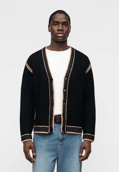 Homme portant un cardigan noir boutonné avec un bord beige, un t-shirt blanc, un jean bleu et une ceinture marron, debout devant un fond uni.