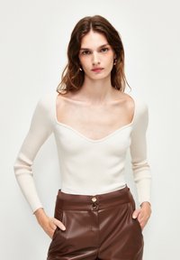 Haut à manches longues côtelé en blanc crème avec un décolleté profond et large. Associé à un pantalon en simili cuir brun taille haute avec des boutons dorés.