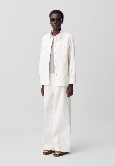 Giubbotto di jeans bianco con bottoni frontali e due tasche sul petto, abbinato a una camicia grigia e pantaloni bianchi a gamba larga. Scarpe con stampa leopardata.