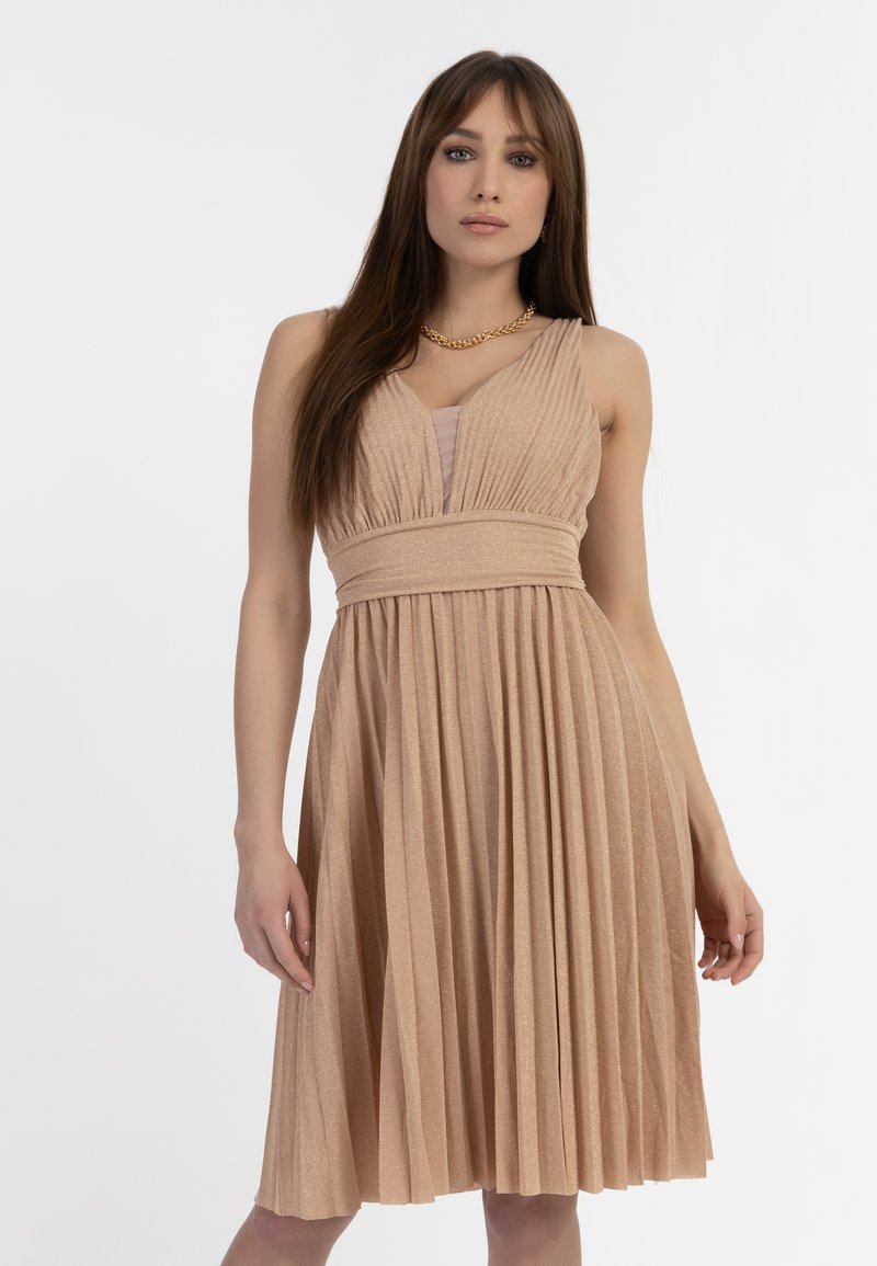 Een beige plissé jurk met een V-hals, getailleerde bovenkant en textiel met structuur. De rok valt onder de knie en toont een glanzende afwerking.