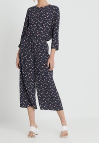 Tuta blu navy con maniche a tre quarti, motivo floreale in rosa, vita elastica e pantaloni larghi. Completata da sandali bianchi con tacco a blocco.
