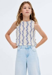Crop top beige clair avec motif floral bleu. Design sans manches, encolure ronde et liens sur les côtés. Associé à un jean taille haute délavé.
