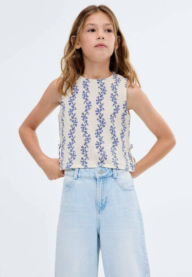 Crop top beige clair avec motif floral bleu. Design sans manches, encolure ronde et liens sur les côtés. Associé à un jean taille haute délavé.