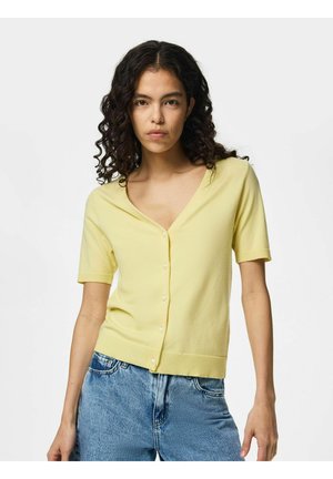 Kvinde med krøllet hår iført en gul kortærmet cardigan med knapper og blå jeans, stående mod en ensfarvet lys baggrund.