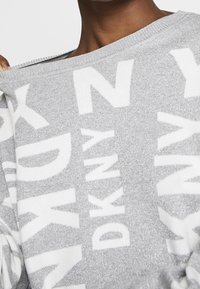 Šedý svetr s lesklým, texturovaným povrchem. Velký bílý potisk "DKNY" a nízko sestřižený výstřih. Měkký pletený materiál, volný střih.