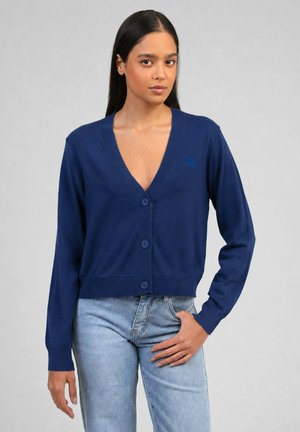 Femme aux longs cheveux foncés portant un cardigan bleu marine boutonné et un jean bleu clair, debout devant un fond gris uni.