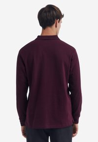 Polo a maniche lunghe di colore bordeaux scuro, materiale morbido, colletto a maglia piatta, lati ventilati, con orlo dritto e design minimalista.