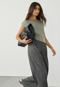 Zielona bawełniana koszulka, dopasowana w ramionach, w zestawie z szarymi spodniami w kratę o szerokich nogawkach. Modelka trzyma czarną skórzaną torbę z złotym akcentem.