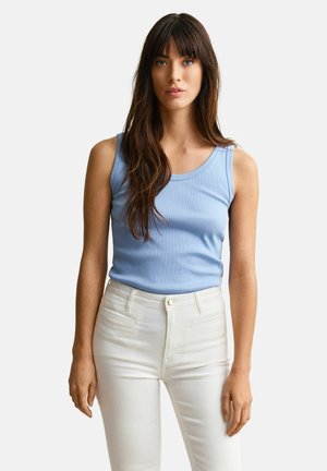Femme aux longs cheveux bruns portant un haut bleu clair sans manches et un pantalon blanc taille haute, debout devant un fond uni.