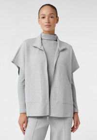 Gilet zip-up grigio con collo alto, maniche corte e due tasche frontali, indossato sopra un maglione grigio a collo alto a coste. Tessuto morbido.