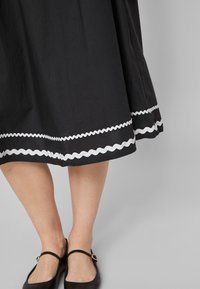 Svart kjol med en utflugen design, med en vit vågig kant vid fållen. Bärs med svarta platta skor, vilket visar benen och vristerna.