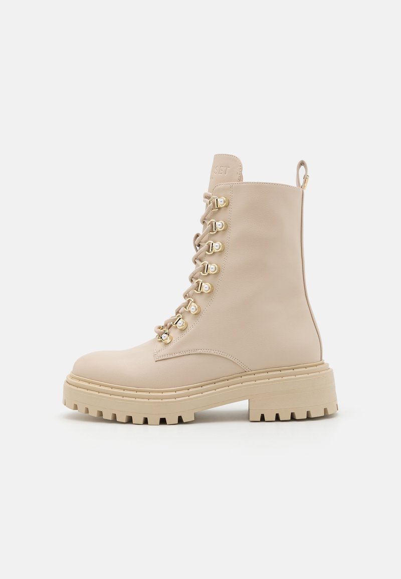 Bottine en cuir beige avec semelle épaisse, œillets dorés, lacets assortis et tirette sur fond blanc.