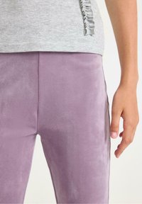 Haut gris clair avec un détail froncé sur le côté, associé à un pantalon lilas slim-fit en tissu doux et lisse. Les pantalons présentent une finition texturée.