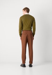 Olivgrüner Strickpullover und braune, maßgeschneiderte Hosen mit umgeschlagenen Säumen. Schwarze Schuhe. Sichtbare Nähte und Gesäßtaschen an den Hosen.