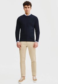 Granatowy sweter z okrągłym dekoltem, pod nim jasna koszula, khaki spodnie slim-fit i beżowe sneakersy. Prosty design z minimalnymi akcentami.