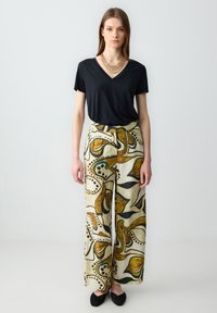 T-shirt noir à col en V associé à un pantalon large avec un motif abstrait jaune et marron, en tissu lisse, et des chaussures plates noires.