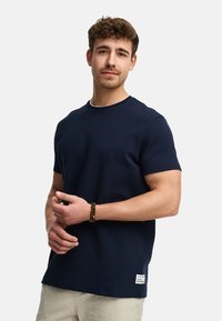 Camiseta de cuello redondo en color navy, fabricada en una mezcla de algodón. Mangas cortas, corte relajado, con una pequeña etiqueta de la marca en el dobladillo. Textura suave.