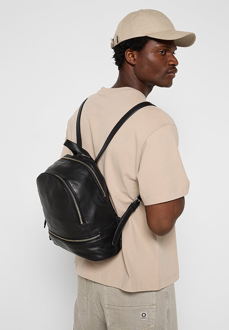 Schwarzer Leder-Rucksack mit gewölbter Form, doppeltem Front-Reißverschluss und verstellbaren Schulterriemen. Wird über einem hellbeigen Hemd getragen.