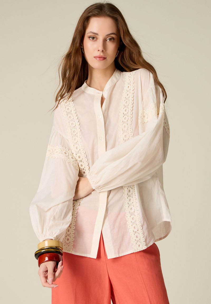 Joven mujer con cabello largo y castaño, que lleva una blusa blanca con detalles de encaje, pantalones color coral y pulseras coloridas apiladas.
