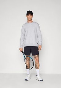 Felpa grigia a maniche lunghe, pantaloncini neri, scarpe da ginnastica bianche, con una racchetta da tennis in mano, tatuaggi sulle gambe e un cappello nero.