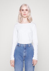 Langärmliges weißes Baumwolloberteil, kombiniert mit hochtaillierten blauen Jeans aus Denim, die über Fronttaschen und einen klassischen Knopfverschluss verfügen.