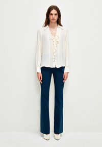 Blusa blanca de manga larga con volantes y detalle de lazo, combinada con pantalones de campana de color azul oscuro y tacones blancos de punta. Fondo liso.