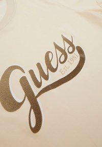 Camiseta cremosa con el logo "Guess" en letras marrones texturizadas, con un estilo cursivo y el detalle "Est. 1981" debajo del nombre de la marca.