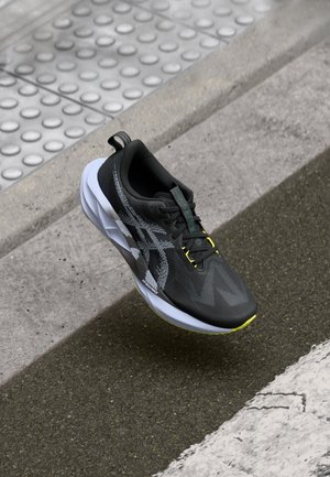 Schwarz-weißer Laufschuh mit gelben Akzenten auf Asphalt nahe einem strukturierten Betonbordstein und taktiler Pflasterung.