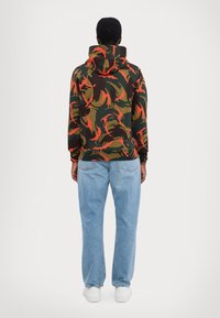 Sudadera con capucha en un patrón de camuflaje multicolor que incluye tonos marrones, verdes y naranja neón, combinada con jeans de color azul claro y zapatillas blancas.