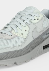 Λευκό και ανοιχτό γκρι αθλητικό παπούτσι Nike Air Max με πάνελ από δίχτυ και δέρμα, δεμένα κορδόνια και ορατή αερόσουλα στη σόλα.