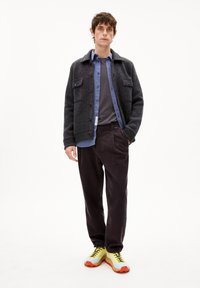 Graue Wolljacke mit zwei vorderen Taschen, über einem blauen Hemd und einem grauen Oberteil getragen. Dunkle Kordhosen und gelbe Sneakers mit roten Sohlen.