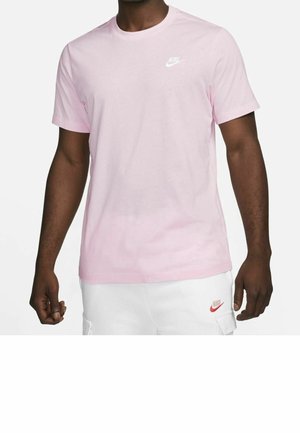 Mann trägt hellrosa Nike-T-Shirt und weiße Nike-Shorts und steht mit entspannten Armen vor weißem Hintergrund.