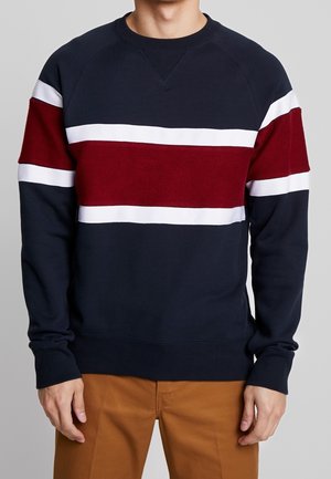 Hombre que lleva una sudadera azul marino con franjas horizontales blancas y rojas a lo largo del pecho y las mangas, combinada con pantalones marrones.