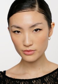 Primo piano di un volto con pelle liscia. Capelli scuri pettinati all'indietro. Le caratteristiche includono sopracciglia definite, eyeliner a ala e piccoli orecchini a cerchio.