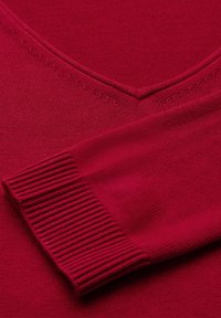 Röd stickad tröja med V-ringning, ribbade manschetter och en mjuk textur. Närbild av tyget visar en slät yta.