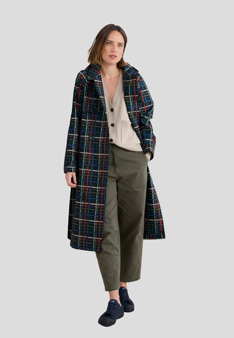 Lungo cappotto scozzese in tonalità blu navy, rosso e verde; presenta grandi bottoni, tasche laterali e una vestibilità rilassata, abbinato a un maglione beige e pantaloni oliva.