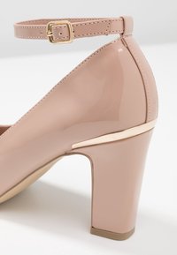 Nude lackläderklack med guldspänne vid vristen och subtil guldig accent nära klacken.