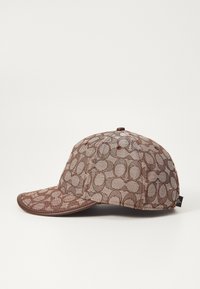 Gorra marrón con un diseño de malla y patrones circulares. La parte frontal tiene un visor rígido, mientras que la parte trasera presenta una etiqueta de tela cerca de la costura.