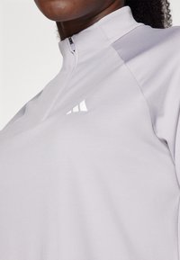 Vaaleanpunertavan violetti vetoketjullinen yläosa, joka on valmistettu pehmeästä kankaasta. Siinä on korkea kaulus ja pieni valkoinen logo rinnassa. Pinta on sileä ja yksityiskohdat ovat vähäiset.