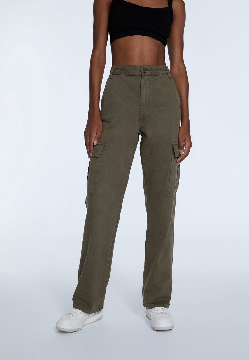 Personne portant un pantalon cargo vert olive, un top court noir et des baskets blanches, debout devant un fond clair uni.