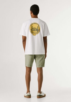 Uomo con maglietta bianca con grafica circolare gialla del Brighton Pier sulla schiena, pantaloncini verde chiaro e scarpe da ginnastica bianche, in piedi di spalle.