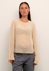 Beige chiffon toppi, jossa on läpinäkyvä muotoilu, koristeltuna pienillä helmimäisillä helmin, pitkissä liehuvissa hihoissa ja pyöreässä kauluksessa.