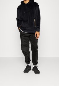 Svart sammetsjacka med dragkedja och snörning på huvan, ihop med svarta manchesterbyxor i cargo-stil och svarta idrottsskor.