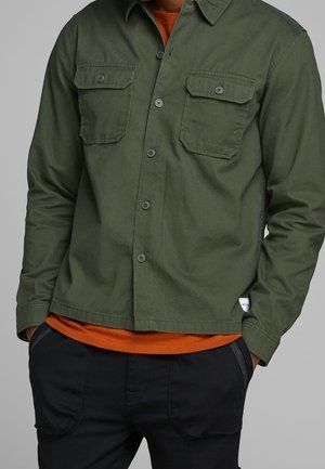 Homme portant une chemise olive à boutons avec des poches sur la poitrine, par-dessus un T-shirt orange et un pantalon noir, les mains dans les poches.