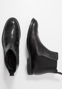 Zwarte leren enkellaarzen met brogue-details, elastische zijkanten, gestructureerde bovenkant en een lage blokhak. Gladde afwerking met een matte uitstraling.